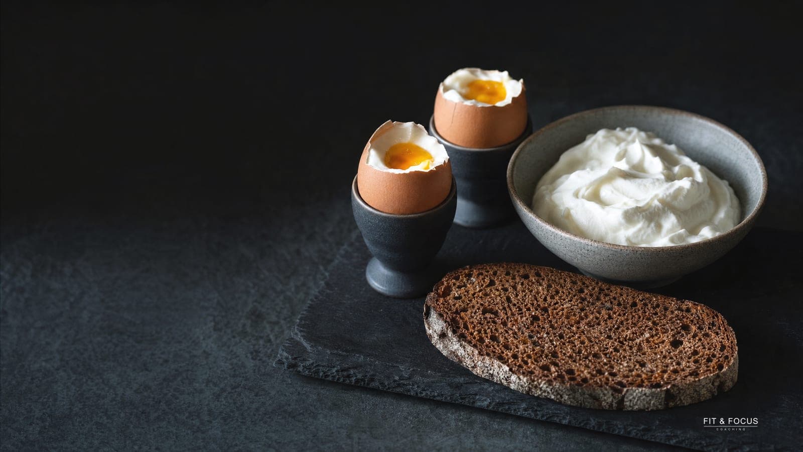 Twee zachtgekookte eieren, een kom yoghurt en een snee roggebrood